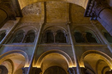 Tarihi bir taştan kilisenin iç kısmının dramatik alçak açılı görüntüsü, sıcak ve çevresel aydınlatma altında devasa sütunlar ve katmanlı kemerler sergiliyor.