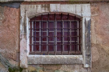 Eski, kavisli bir pencerenin yakın çekimi paslanmış demir parmaklıklar yıpranmış taş bir duvara yerleştirilmiş, kırmızı renkli camları grafiti ile kaplanmış.