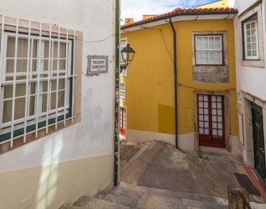 Renkli binalar arasında dar, güneşli bir ara sokak rüzgarı. Sarı bir cephe, kırmızı bir kapı ve fayanslı bir sokak tabelası var.