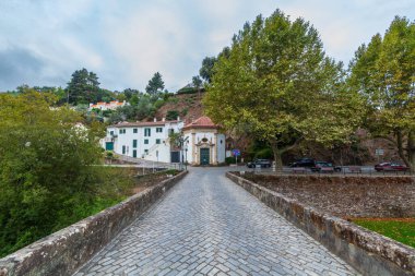 Bir kaldırım taşı köprüsü, bulutlu bir gökyüzünün altındaki yamaçtaki yemyeşil ağaçların arasına yerleştirilmiş kırmızı kiremitli çatısı olan beyaz bir şapele doğru gidiyor.