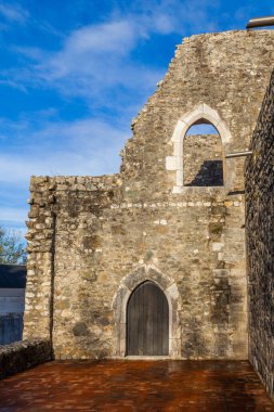 Leiria. Antik bir taş harabesinin güneşli cephesinde karanlık bir ahşap kapı ve parlak mavi bir gökyüzüne kurulmuş kemerli bir pencere, sıcak terasotta fayans zemini ile tarihi bir sükunet ve mimari detay sahnesi yaratıyor.