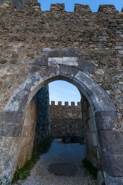Leiria. Düşük açılı bir bakış açısı, yıpranmış taştan bir kemer yolundan geçerek, eski bir kaleye giden yolu ortaya çıkarır, berrak mavi gökyüzüne karşı yükselen, tarihi bir ihtişam ve gizem hissi uyandıran,