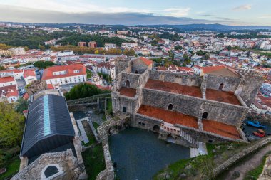 Leiria. Bir kale avlusundan geniş bir hava manzarası, antik taş duvarlarını ve terracotta çatılarını gözler önüne seriyor, kırmızı kiremitli binalardan oluşan canlı bir şehir manzarası, dramatik bulutlu gökyüzünün altındaki yeşil tepelerin arasına yerleşmiş..