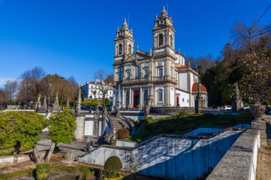 Braga 'daki bir Bom Jess de Monte süslemeli taş kilise. İkiz çan kuleleri açık mavi gökyüzünün altında görkemli bir şekilde duruyor. Beyaz cephesi ve kırmızı kapıları ziyaretçilerin gezindiği ön plandaki büyük bir heykel ile çelişiyor..