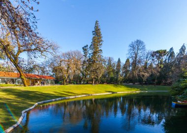Sakin bir park sahnesinde mavi gökyüzünü ve çevreleyen ağaçları yansıtan sakin bir göl, çimenli bir kıyısı, kırmızı kiremitli bir çatısı olan bir bina ve güneşli günün keyfini çıkaran insanlar yer alıyor.