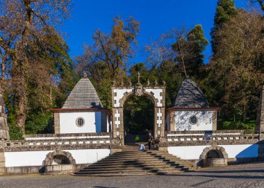 Tepesinde haç bulunan büyük taş kemer yolu, bir merdivene giriş görevi görüyor. İki küçük şapelin yan tarafında konik çatılar var. Hepsi de ağaçların ve açık mavi gökyüzünün önünde.