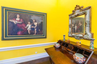 Canlı sarı bir odada, renkli porselen vazolar ve heykelciklerle süslenmiş koyu ahşap bir konsol masasının üzerindeki süslü aynada yansıtılan, yaldızlı bir çerçevedeki figürlerin klasik bir resmi sergilenmektedir..