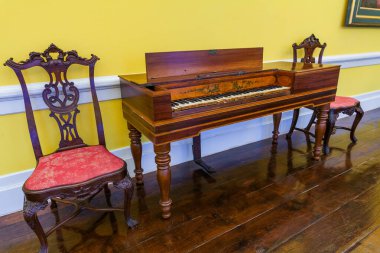 Süslü ahşap işlemeli ve çiçek işlemeli zarif bir antika klavsen kırmızı döşemeli uyumlu bir sandalyenin yanında duruyor, parlak sarı bir duvara karşı parlak, koyu ahşap bir zeminde sergileniyor..