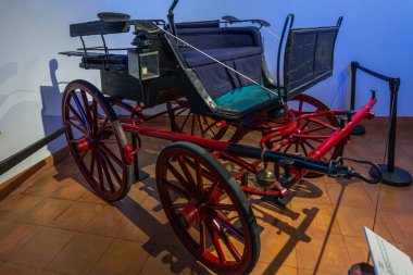 Biscainhos Müzesi. - Braga. Büyük ahşap tekerlekleri, siyah gövdesi ve katlanmış üstü, turkuaz koltuk minderleri, yan feneri ve loş ışıklı müze ortamında bariyerin arkasında sergilenen koşum direği olan klasik kırmızı fageton at arabası..