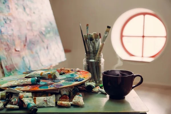 Le lieu de travail de l'artiste, pinceaux, peintures, toile sur le chevalet. Une tasse avec une boisson chaude. Mise au point sélective, image tonique . — Photo
