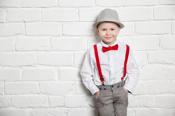 Kleiner Junge mit roter Fliege, Hosenträgern und weißem Hemd vor einer weißen Backsteinwand. — Stockfoto