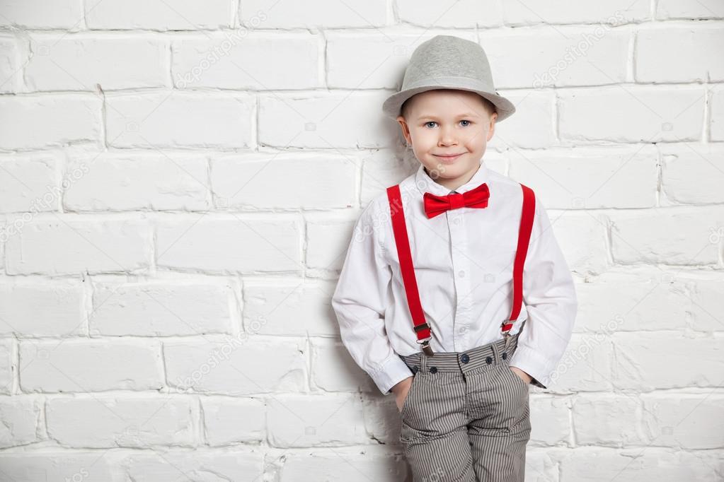 little boy wearing a red bow tie, suspenders and white shirtand against a white brick wall.