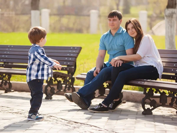 若い親と幼い息子公園で。父と母、公園のベンチに座っていると、彼の息子を見て. — ストック写真