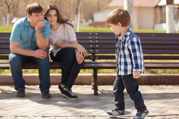 若い親と幼い息子公園で。父と母、公園のベンチに座っていると、彼の息子を見て. — ストック写真