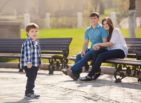 Padres jóvenes e hijo pequeño en el parque. Padre y madre sentados en un banco en el parque y mirando a su hijo . — Foto de Stock