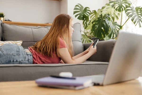 Relajado sonriente joven mujer mensajes de texto en el teléfono celular tumbado en el sofá en casa, vista lateral — Foto de Stock