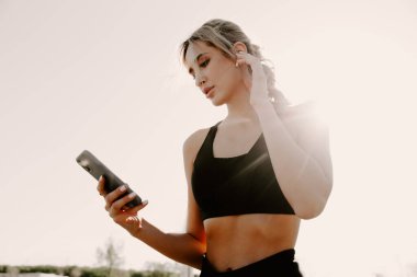 Kulaklıklı genç bir kadın şehirde koşuyor ve cep telefonu arıyor..