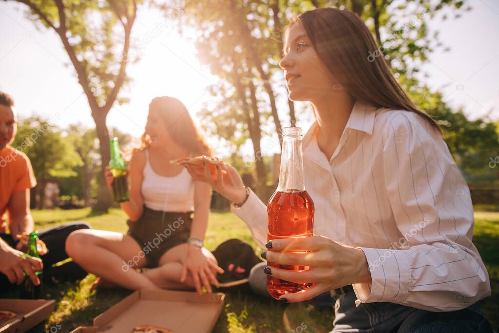 Young girl drinking alcohol and eating pizza with her friends outdoors, background image, focus on foreground.