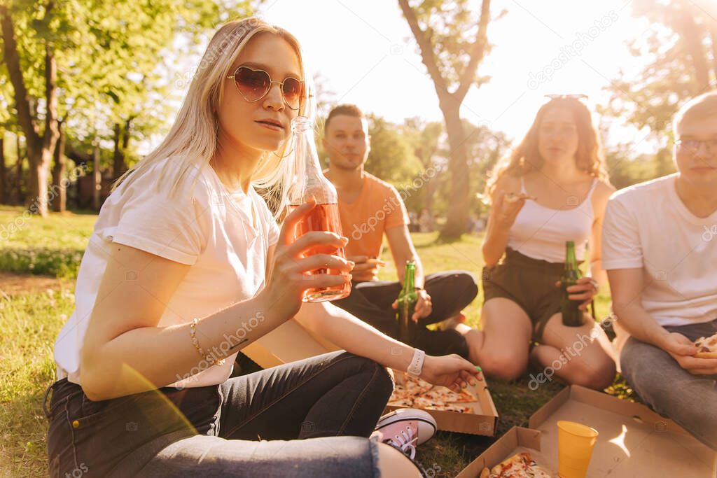 Young blonde girl in sunglassesdrinking alcohol and eating pizza with her friends outdoors.