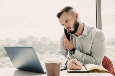 Genç adam dizüstü bilgisayarıyla ilgileniyor, cep telefonuyla konuşuyor ve notlar alıyor..