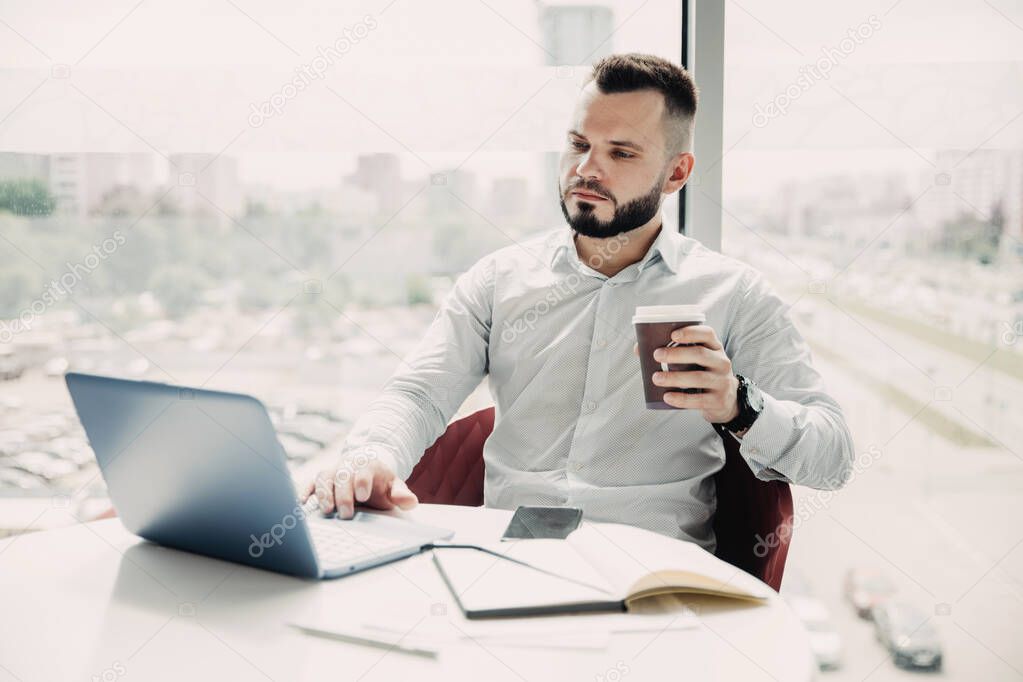 Young bearded businessman working in office on laptop and drinking coffee from paper cup.