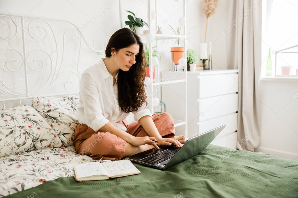 Woman sitting on bed working and using laptop. Freelancer, work from home.