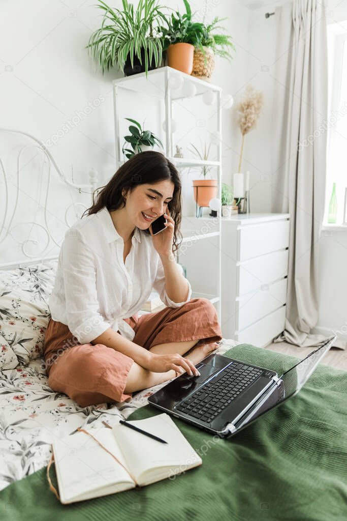 Woman sitting on bed talking on the phone and working using laptop.