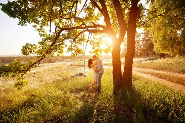 Jovem casal beijando sob grande árvore com balanço ao pôr do sol — Fotografia de Stock