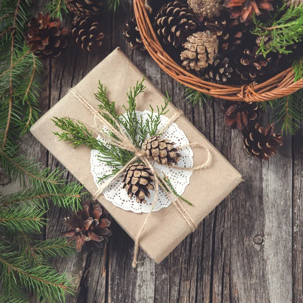 Hand crafted gift on rustic wooden background and a basket with fir branches and cones — Stock Photo, Image