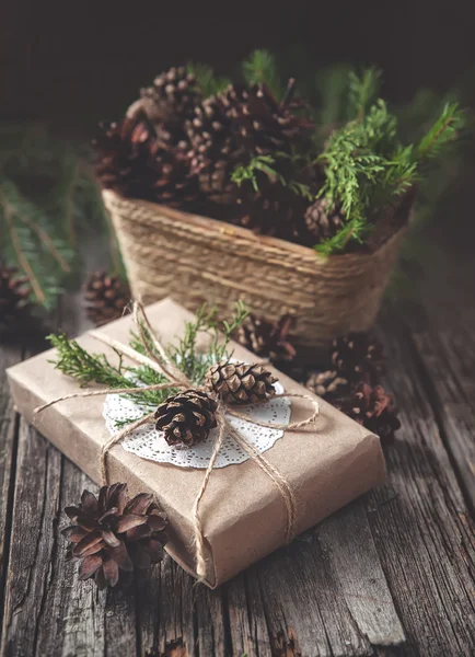 Hand crafted gift on rustic wooden background and a basket with fir branches and cones — Stock Photo, Image