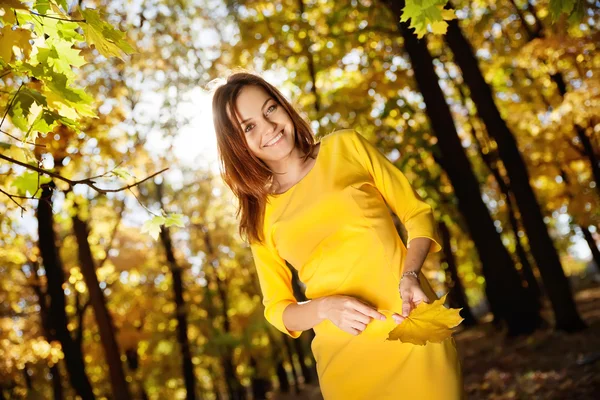 Молодая женщина в желтом платье с осенними листьями в руке и осенью желтый кленовый сад фоне — стоковое фото