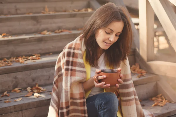 Young woman resting and drinking tea sitting in autumn garden on the steps, wrapped in a woolen plaid blanket. — Stock Photo, Image