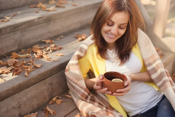 Jonge vrouw rusten en het drinken van thee zitten in herfst tuin op de trappen, verpakt in een wollen geruite deken. — Stockfoto