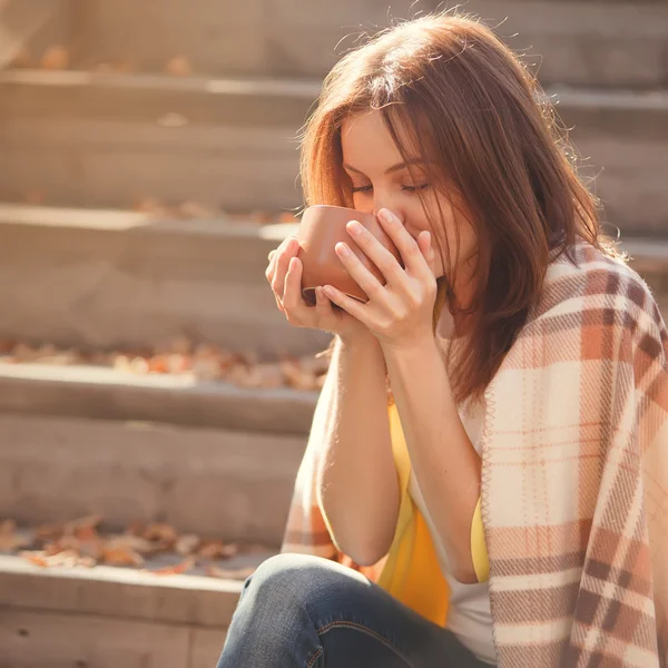 Giovane donna che riposa e beve tè seduta nel giardino autunnale sui gradini, avvolta in una coperta a quadri di lana . — Foto Stock