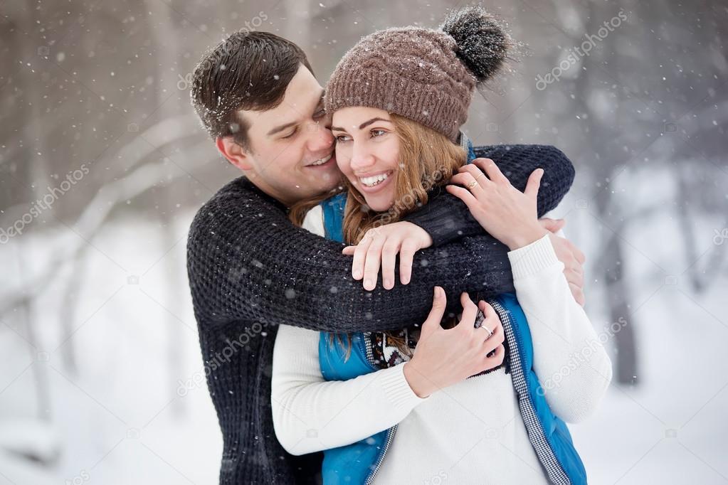 Young couple in love walking in winter park. It's snowing, winter. Young man embraces the girl. She laughs.