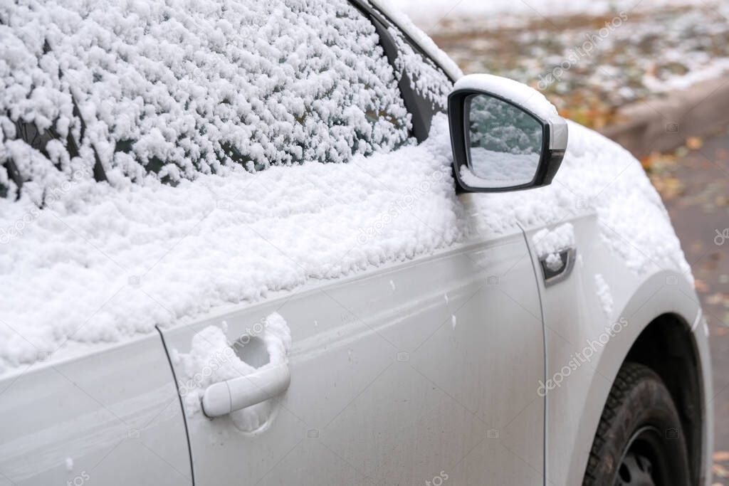 White passenger car is parked near a residential building in the city covered with the first autumn snow close-up fragment