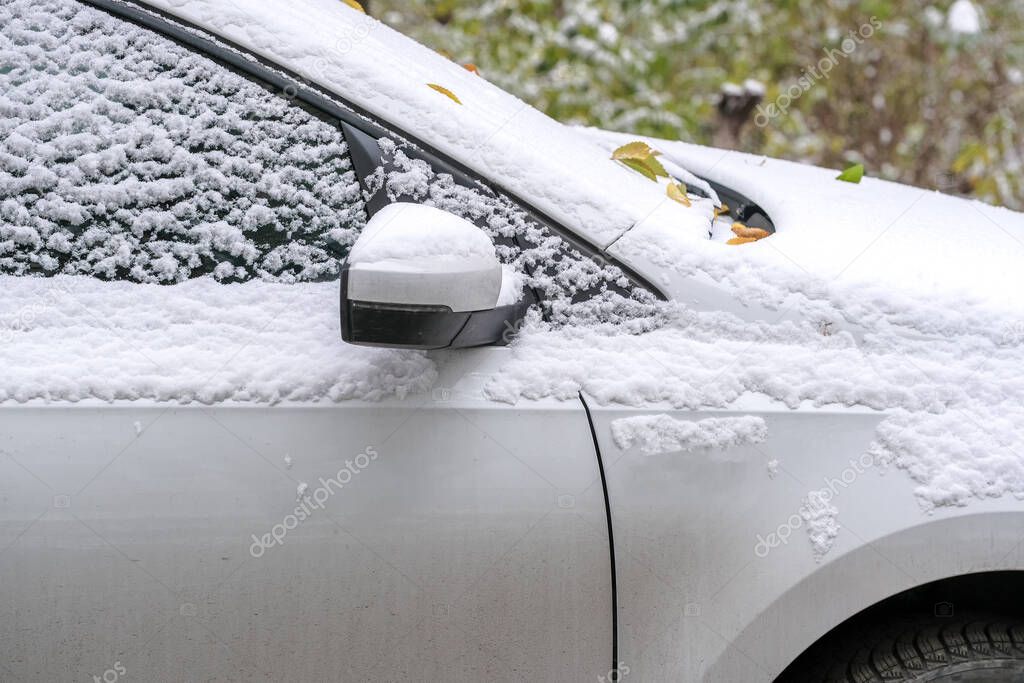 White passenger car is parked near a residential building in the city covered with the first autumn snow close-up fragment