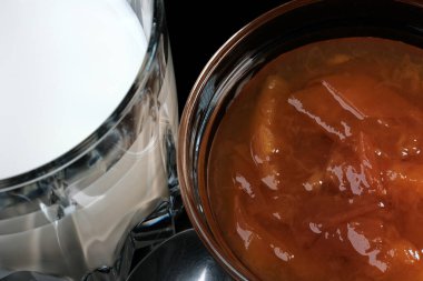 Şeftali reçelli şeffaf cam bir kase siyah arka planda bir bardak sütün yanında duruyor.