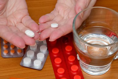 Bir bardak temiz su ve hap paketinin yanında iki el beyaz hap sergiliyor..