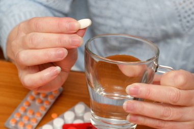 Eller bir hapı temiz bir bardak suyun üzerinde, tahta bir masanın üzerinde tutar..