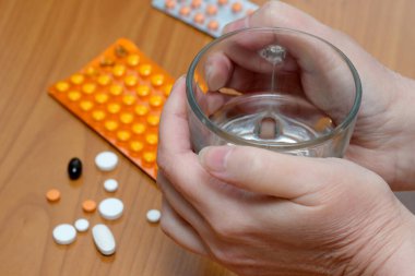 Elleri bir bardak suyu ahşap bir yüzeyde renkli hapların yanında tutuyor..