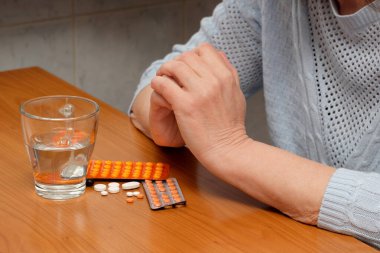 Bir insan, elinde ilaç ve bir bardak suyla ahşap bir masada düşünceli bir şekilde oturur..