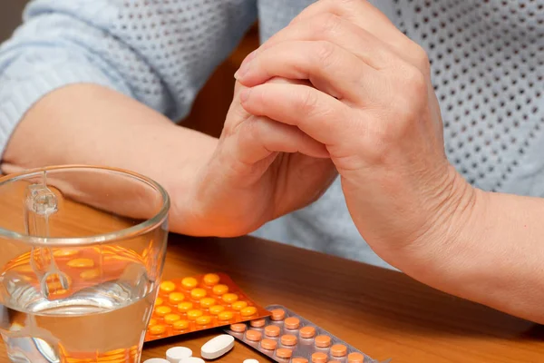 Yaşlı bir yetişkinin elleri ilaç ve su dolu bir masada dinleniyor..