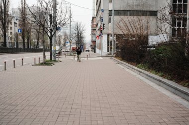 Issız kaldırım boyunca gri bir şehir manzarasında tek başına yürüyen bir figür. Shevchenko Bulvarı. Kyiv, Ukrayna. 16 Aralık 2025.