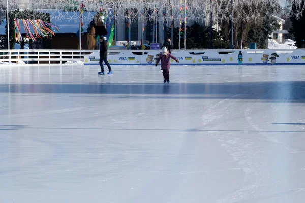 Bir çocuk buzun üzerinde mutlu bir şekilde kayarken bir yetişkin parlak güneş ışığında yakınlarda süzülüyor. Kyiv, Ukrayna. 21 Şubat 2026. Ukrayna Ulusal Kompleks Expocenter, Maslenitsa