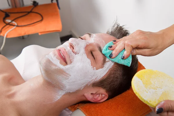 Terapeuta aplicando uma máscara facial a um belo jovem em um spa — Fotografia de Stock