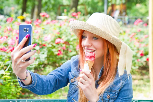 Красивая женщина с рыжими волосами ест вкусное мороженое, делая селфи. Девушка отдыхает в парке — стоковое фото