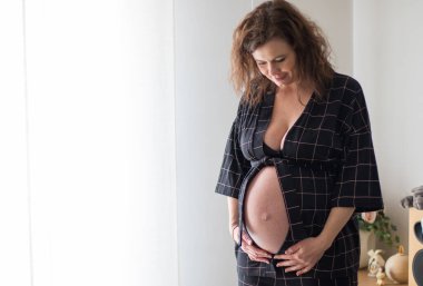 Genç, güzel, hamile bir kadın pencerenin yanında duruyor ve tatlı tatlı karnına dokunuyor.