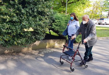 Genç bakıcı yaşlı bir beyefendiye parkta yürümesine yardım ederken eşlik ediyor. 
