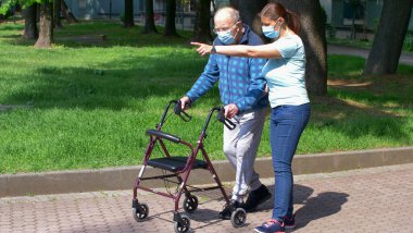 Genç bakıcı yaşlı bir beyefendiye parkta yürümesine yardım ederken eşlik ediyor. 